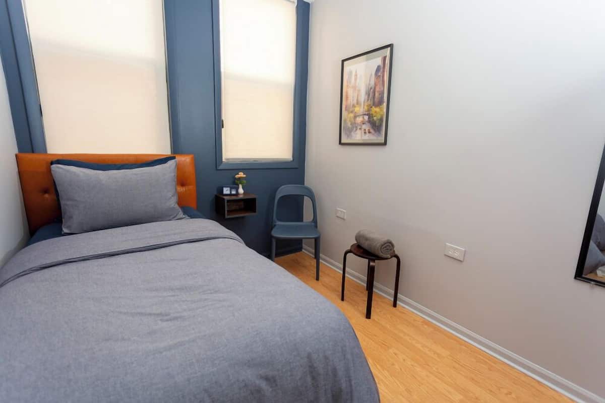 A bedroom with a bed, a chair, a small table with a lamp, and a painting on the wall.