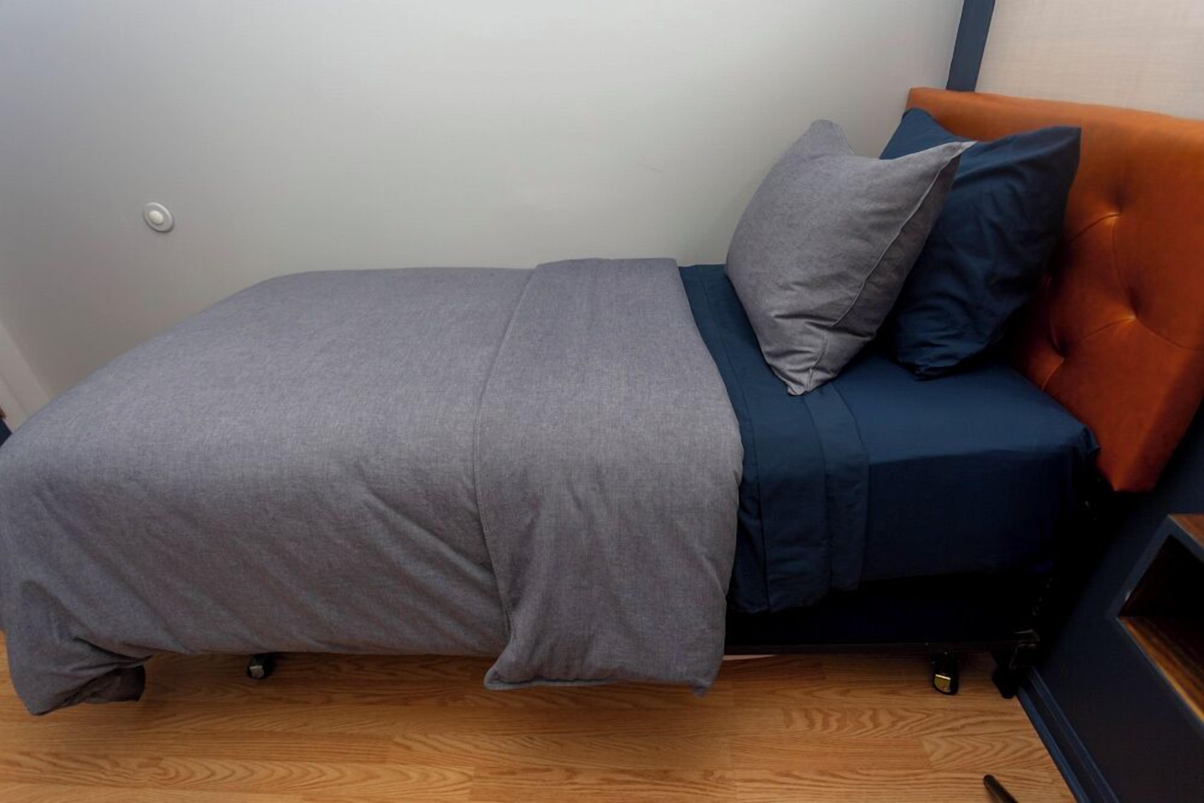 Una stanza piccola con un letto singolo, copripiumino grigio, materasso blu e testiera arancione.