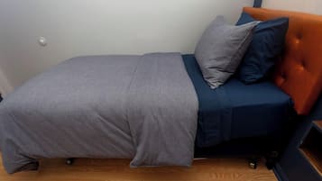 Una stanza piccola con un letto singolo, copripiumino grigio, materasso blu e testiera arancione.