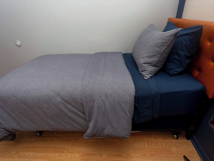 Una stanza piccola con un letto singolo, copripiumino grigio, materasso blu e testiera arancione.