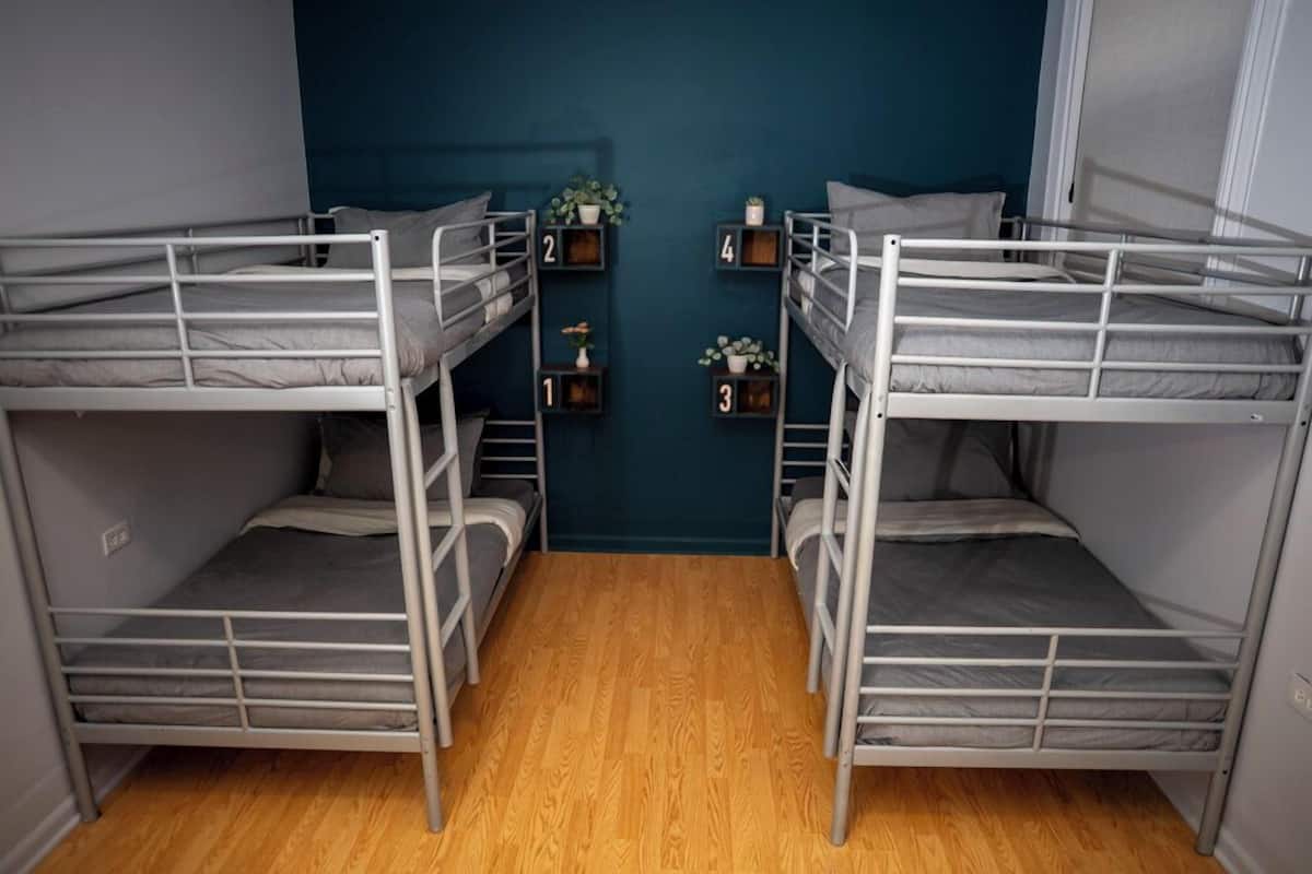 A room with four bunk beds arranged in a square pattern, wooden flooring, and a teal-colored wall with small shelves holding decorative items.