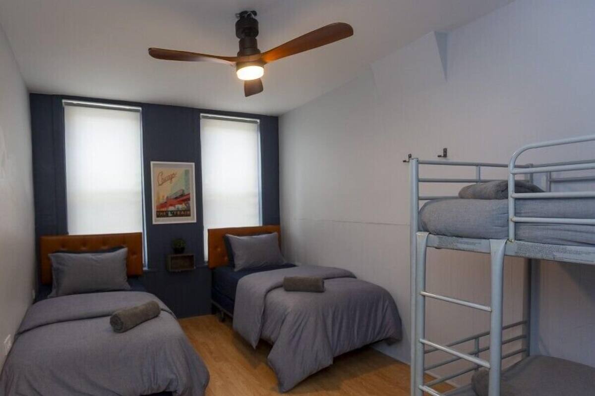 A compact room with two single beds, a bunk bed, a ceiling fan, and two windows with blinds.