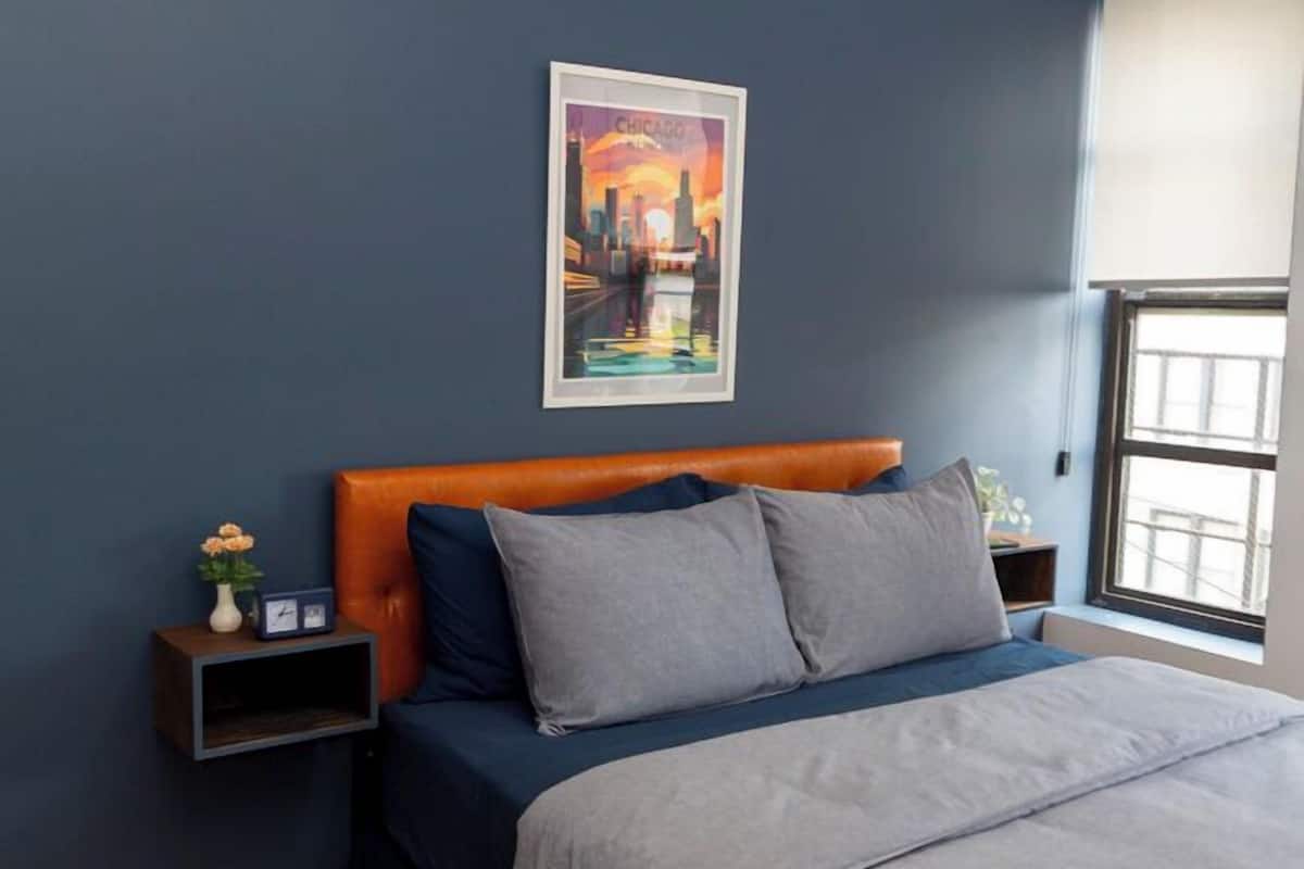 A bedroom with a bed, a nightstand, a ceiling fan, a window, and a painting on the wall.