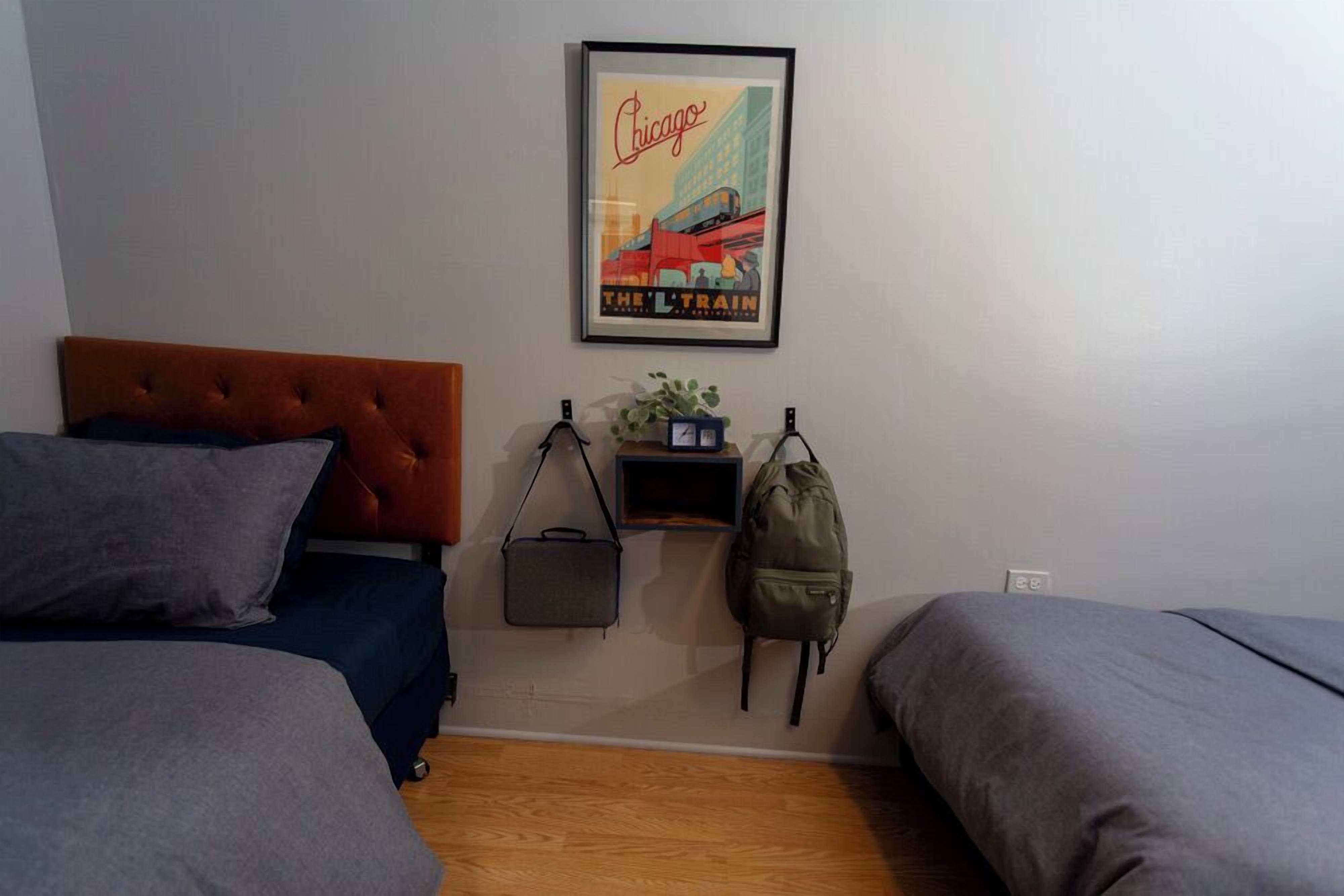 Ein Schlafzimmer mit zwei Betten, einer Kopfteil, einem gerahmten Bild an der Wand, einer kleinen Ablage mit einer Pflanze und zwei Taschen sowie einem Holzboden.