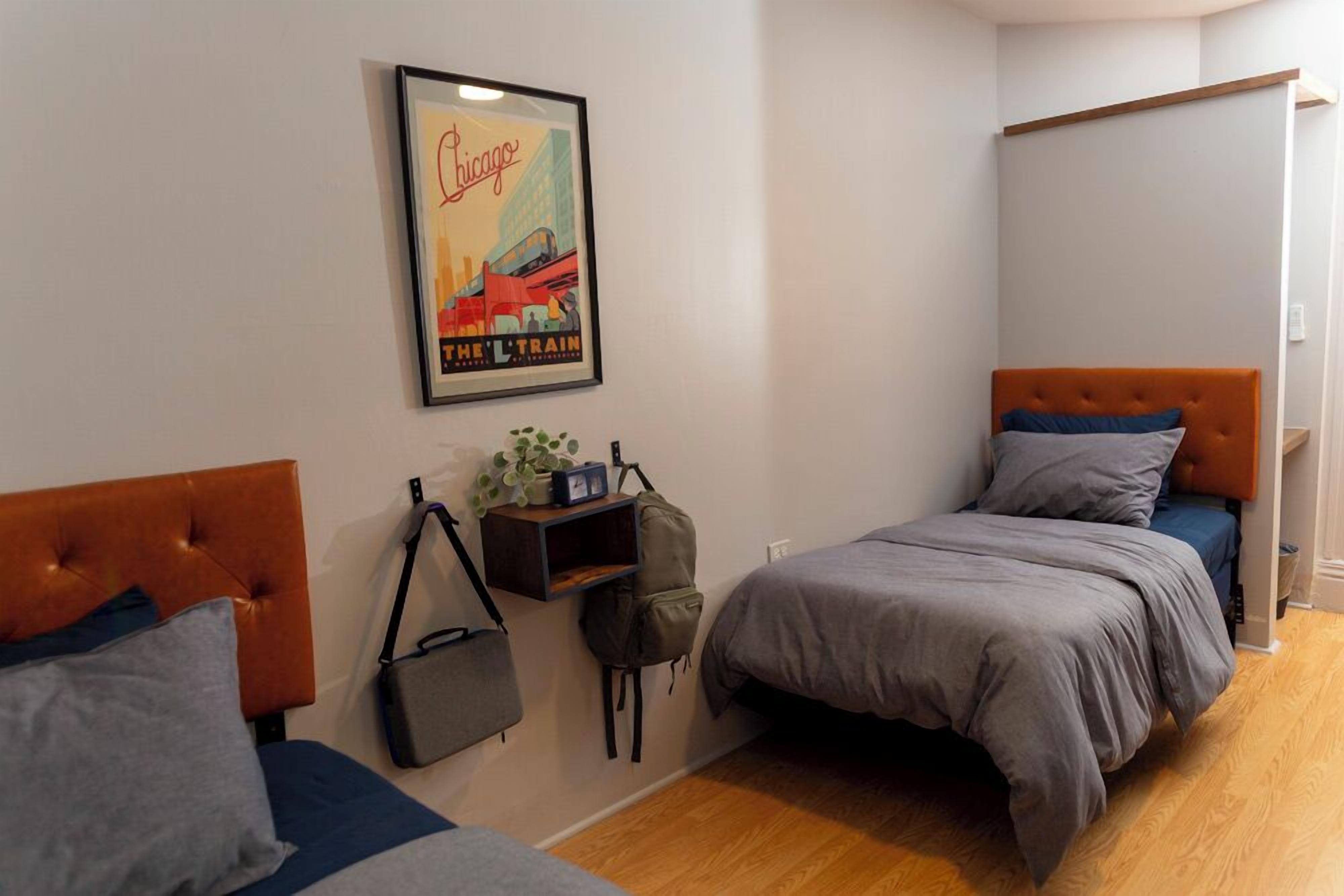 A bedroom with two beds, a framed picture of Chicago on the wall, and a small shelf with a plant and a bag.
