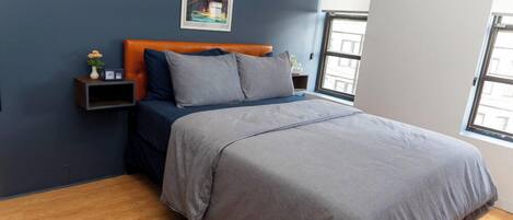 A bedroom with a bed, a nightstand, a wall clock, a framed picture, and a window.