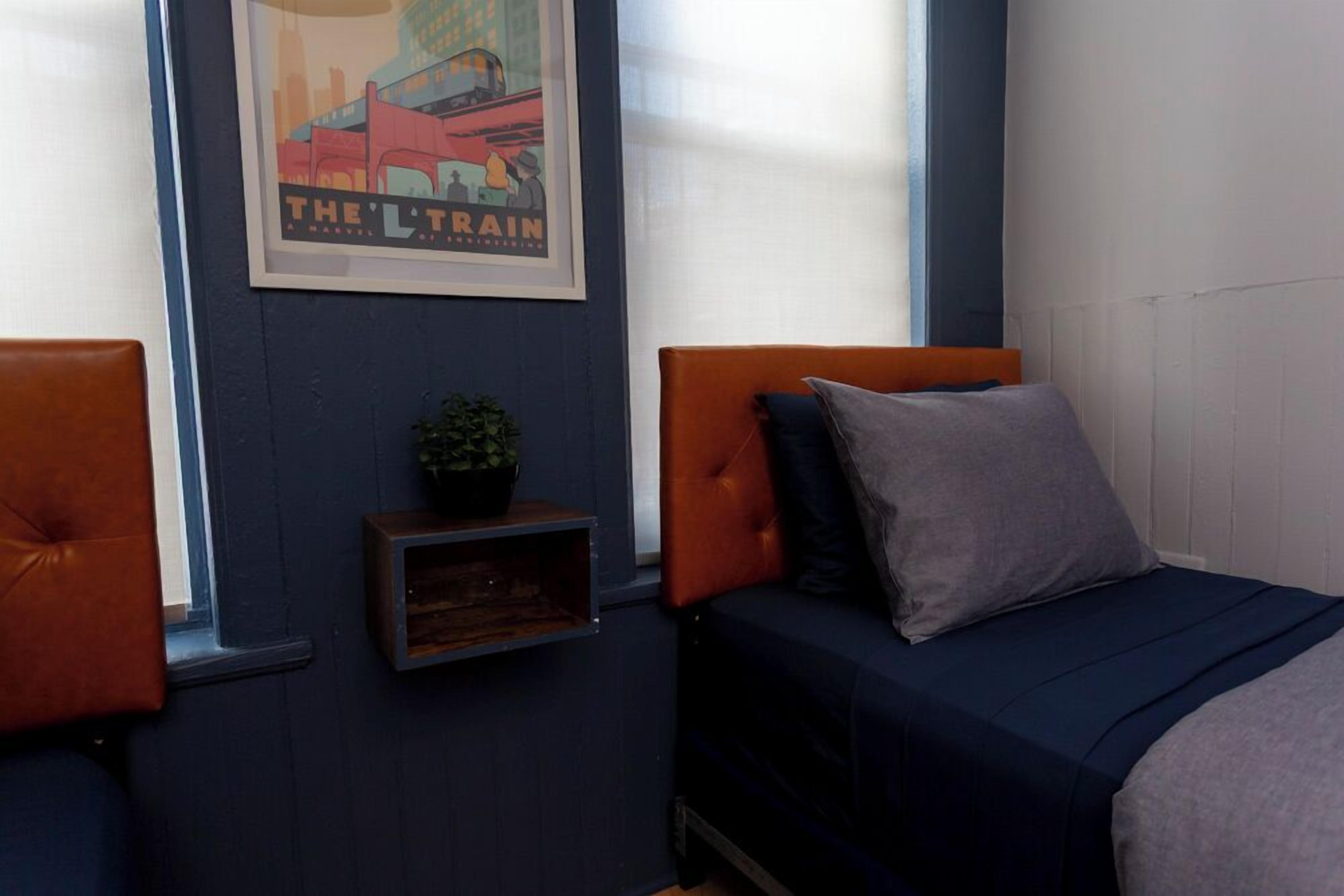 Une chambre avec deux lits, l’un doté d’une tête de lit orange et l’autre d’une tête de lit grise. Un cadre avec une affiche est accroché au mur, et une petite étagère supporte une plante.