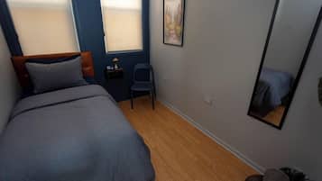 Quarto com cama, uma cadeira, mesa pequena, espelho e janela.