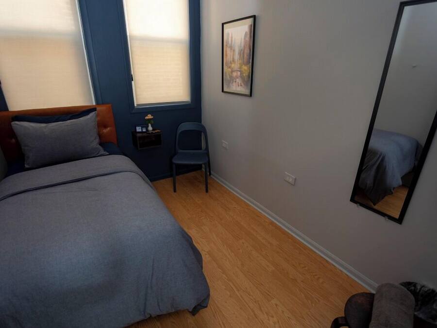 Quarto com cama, uma cadeira, mesa pequena, espelho e janela.