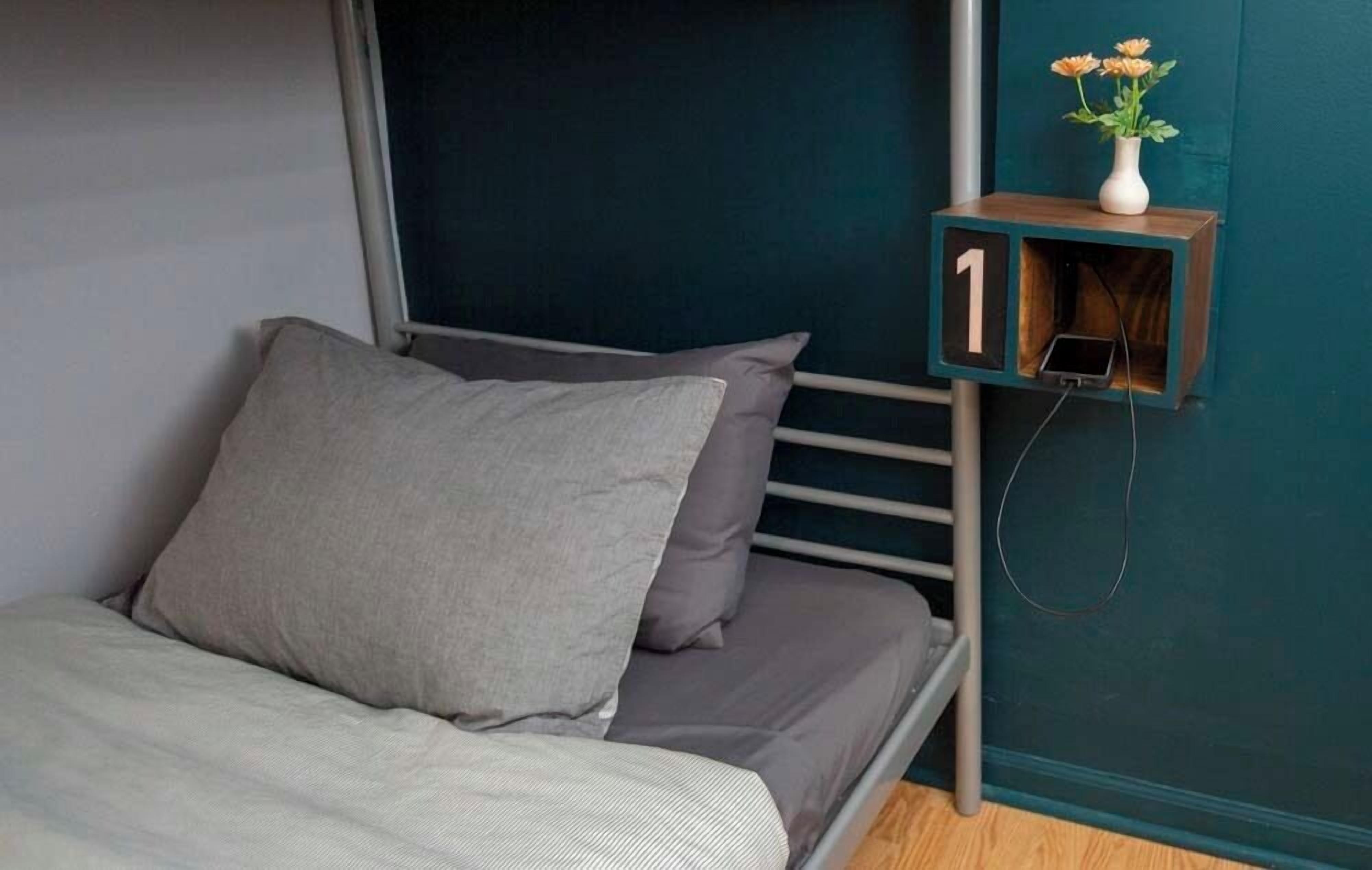 A bed with grey bedding, a small bedside table with a phone, and a wall-mounted shelf with a vase of flowers.
