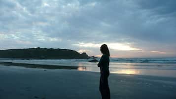 A person standing on a beach with a view of the ocean and a distant landmass under a cloudy sky.