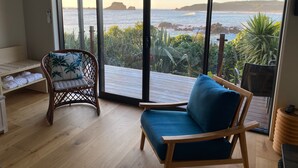 A balcony with a wooden floor, a wicker chair with a cushion, a blue cushioned chair, and a stack of towels.