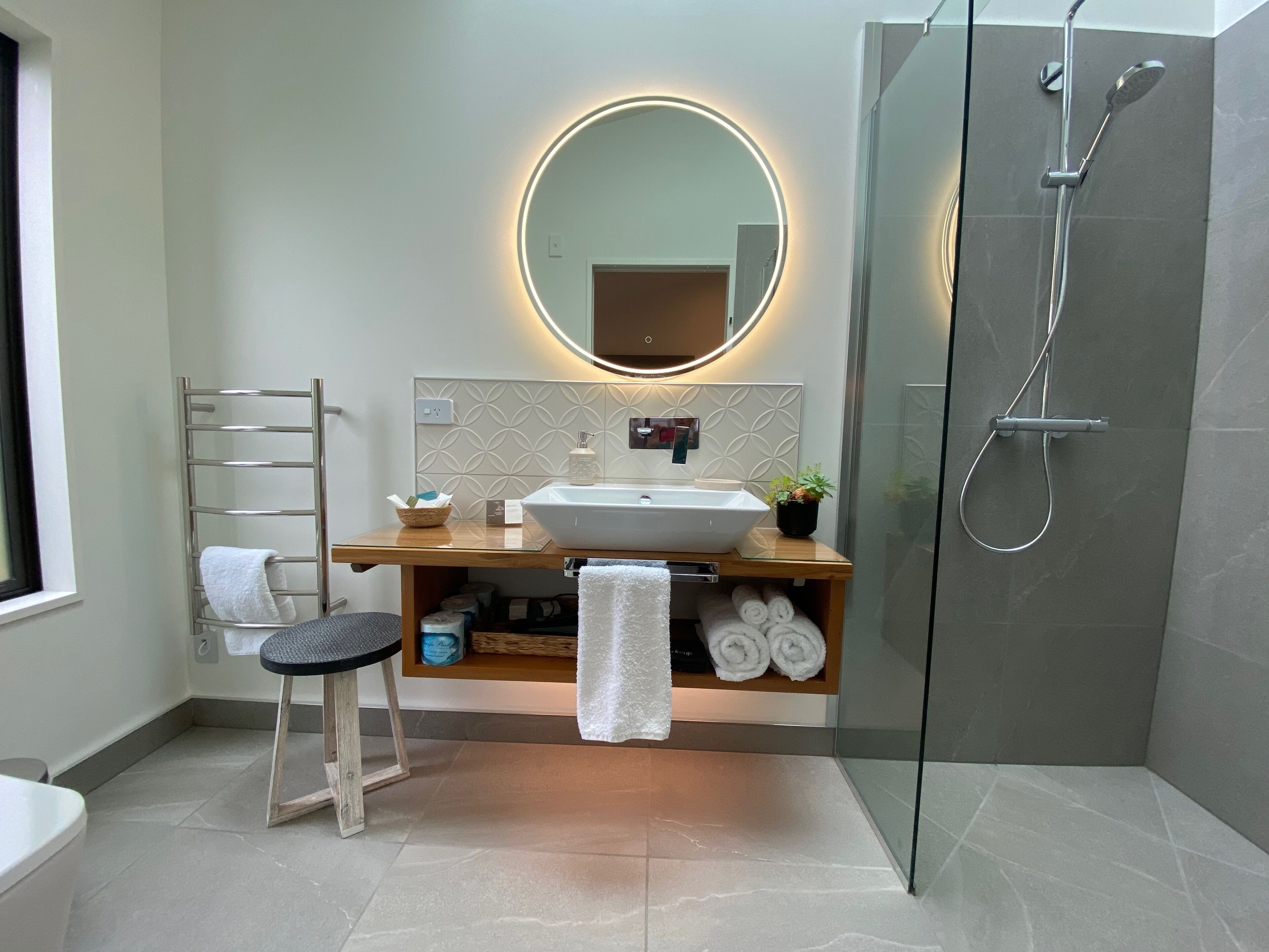 A modern bathroom with a glass-enclosed shower, a wooden vanity with a round mirror, and a towel rack.