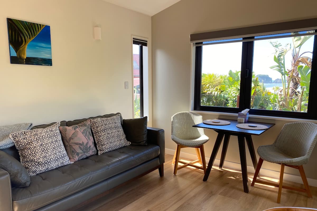 Ein modernes Wohnzimmer mit einem Ledersofa, zwei Korbstühlen, einem kleinen Holztisch und einem großen Fenster mit Blick auf Grünflächen und ein Gewässer.