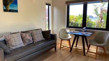A modern living room with a leather sofa, two wicker chairs, a small wooden table, and a large window offering a view of greenery and a body of water.