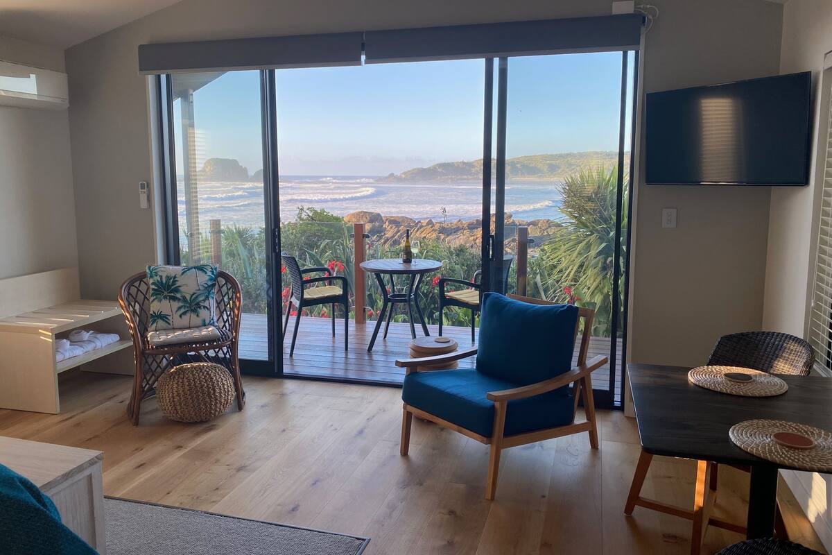 Ein modernes Wohnzimmer mit einer großen Schiebeglasdoor zum Balkon mit Meerblick. Zwei Sessel, ein kleiner Tisch und ein Sofa sind vorhanden.