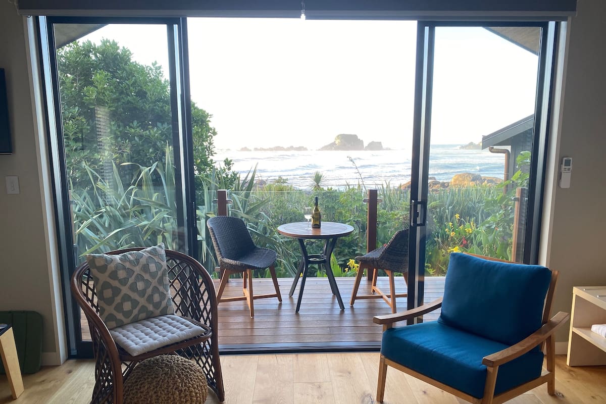 Ein Zimmer mit einer großen Schiebetür und einem Balkon mit Meerblick. Auf dem Balkon befinden sich zwei Korbstühle und ein blaues Sofa.