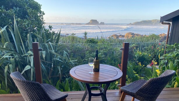 A wooden deck with two wicker chairs and a round table, overlooking a rocky coastline and lush greenery.