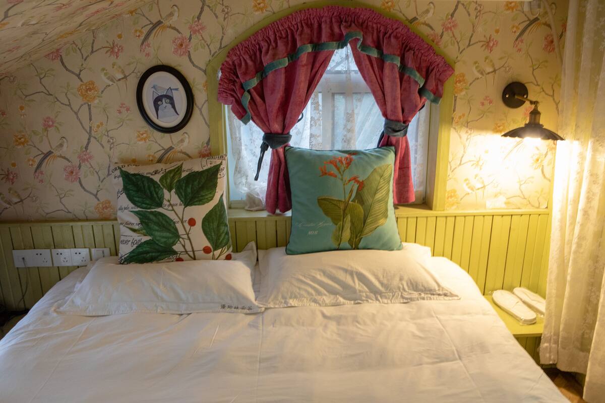 A bedroom with floral wallpaper, a bed with white bedding, a window with a pink curtain, and a bedside lamp.