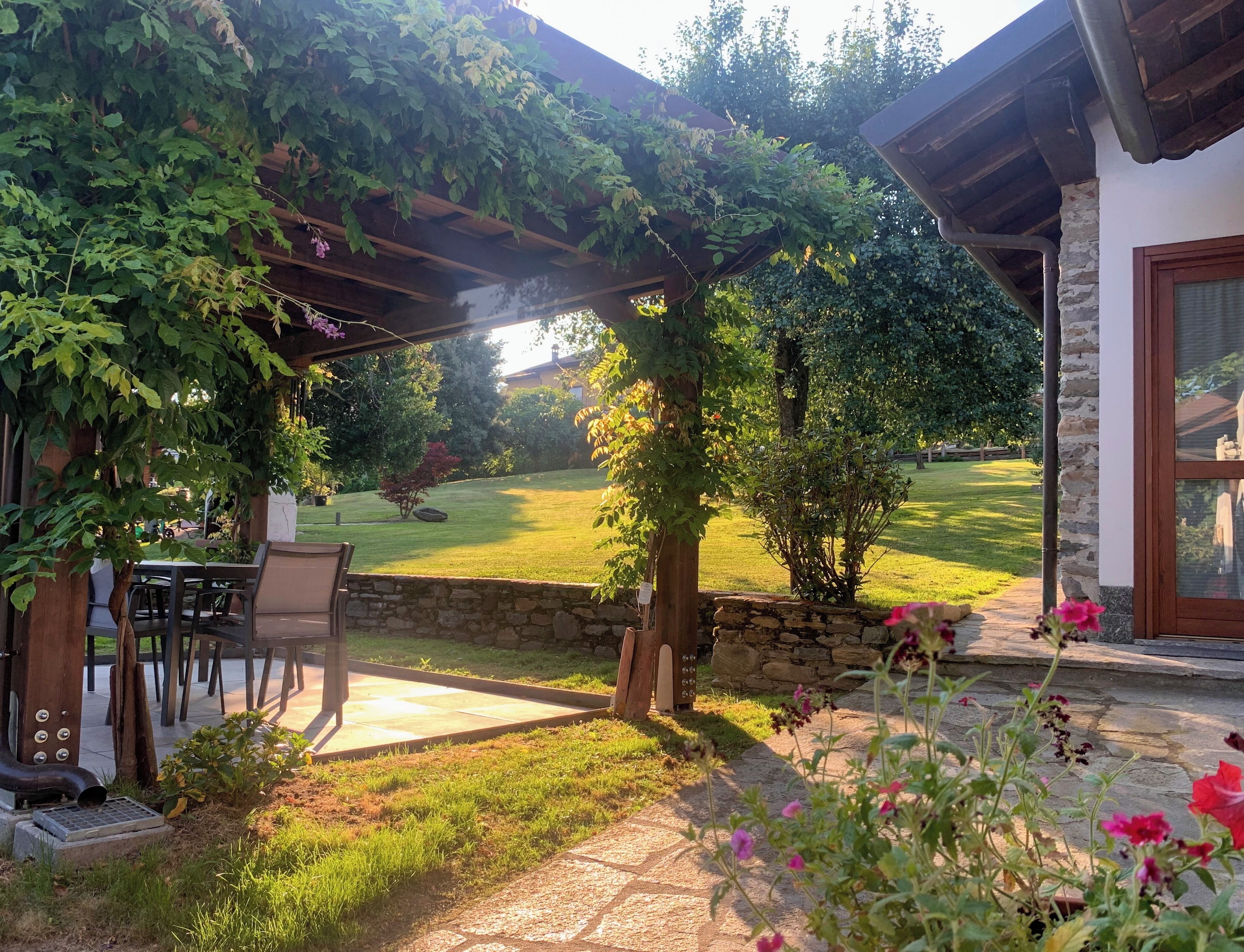 Un patio con pérgola cubierta de vegetación, un muro de piedra y vistas a un césped y árboles bien cuidados.