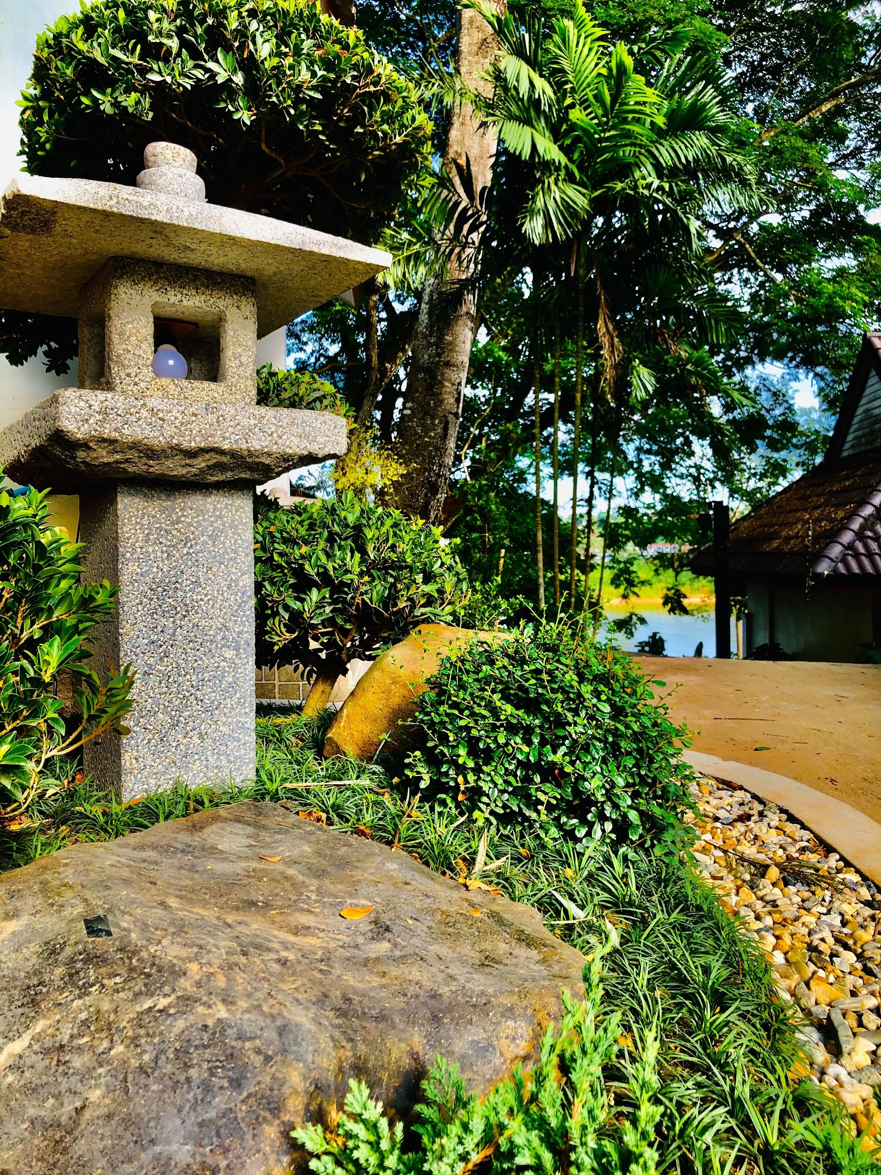 Un farol de piedra rodeado de abundante vegetación y una estructura con techo de paja en un jardín.