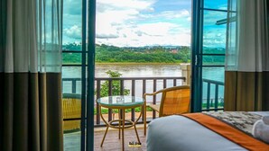 Habitación con un ventanal grande que da a un río y vegetación. Cuenta con una cama con cabecera blanca y naranja, una mesita redonda pequeña y dos sillas de mimbre.