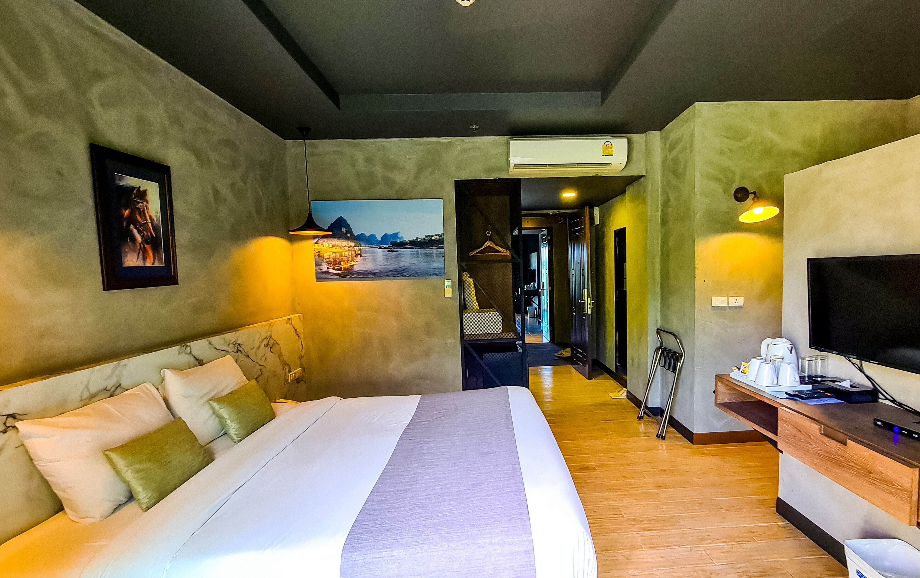 A hotel room with a large bed, a desk with a flat-screen TV, an air conditioning unit, and a hallway leading to other rooms.