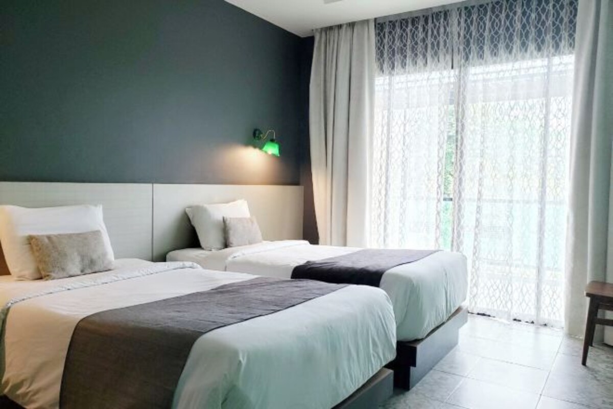 Une chambre d’hôtel avec deux lits, une table de chevet, une lampe et une grande fenêtre avec des rideaux légers.