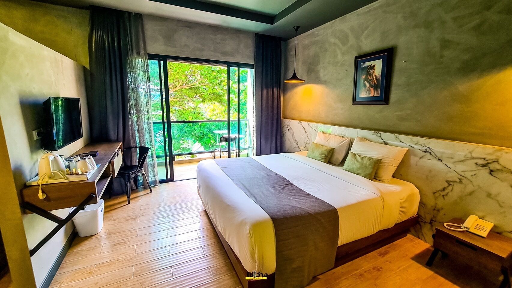 A hotel room with a large bed, a desk with a chair, a TV, and a balcony with a view of greenery.