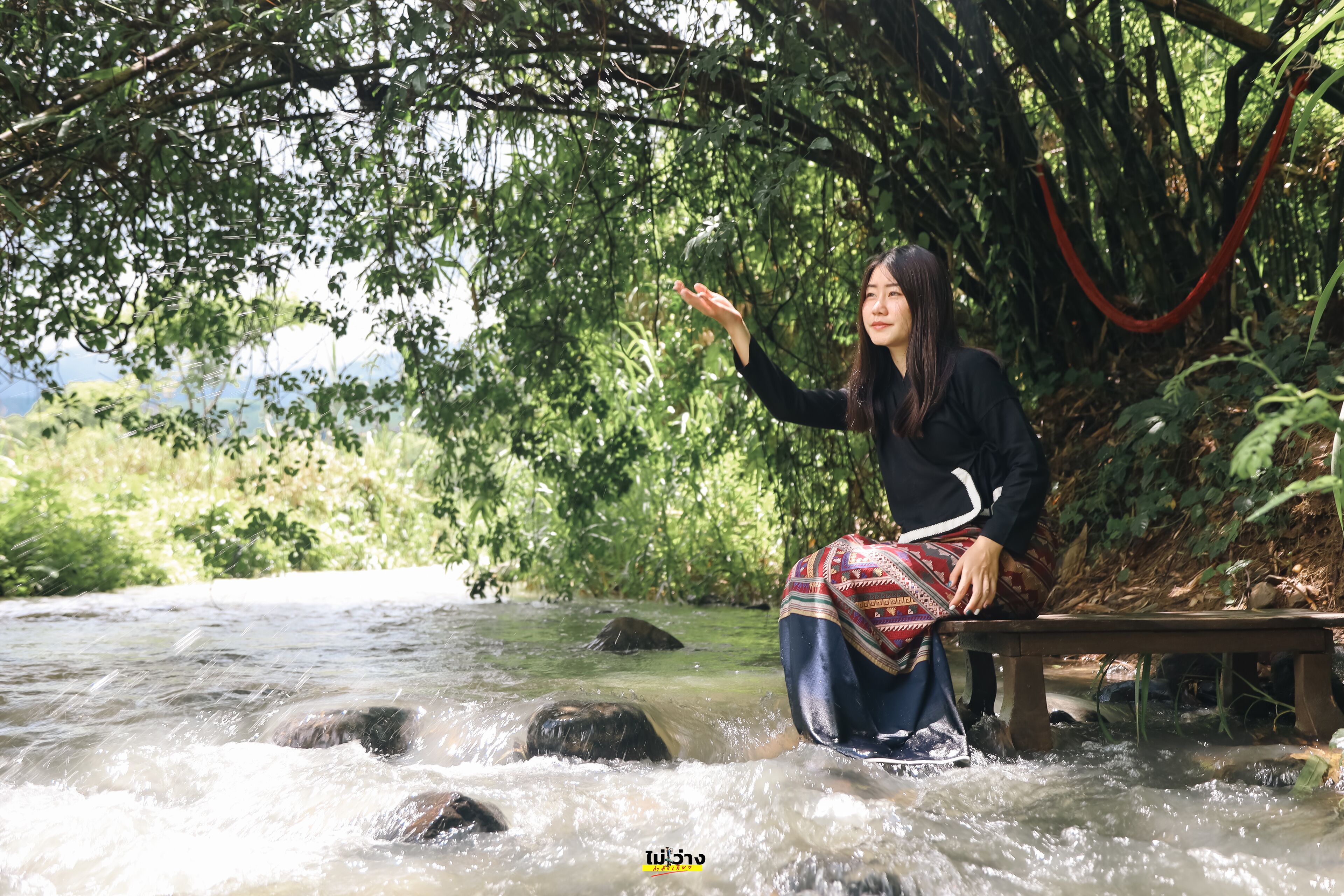 A person sitting by a river with rocks and greenery in the background.