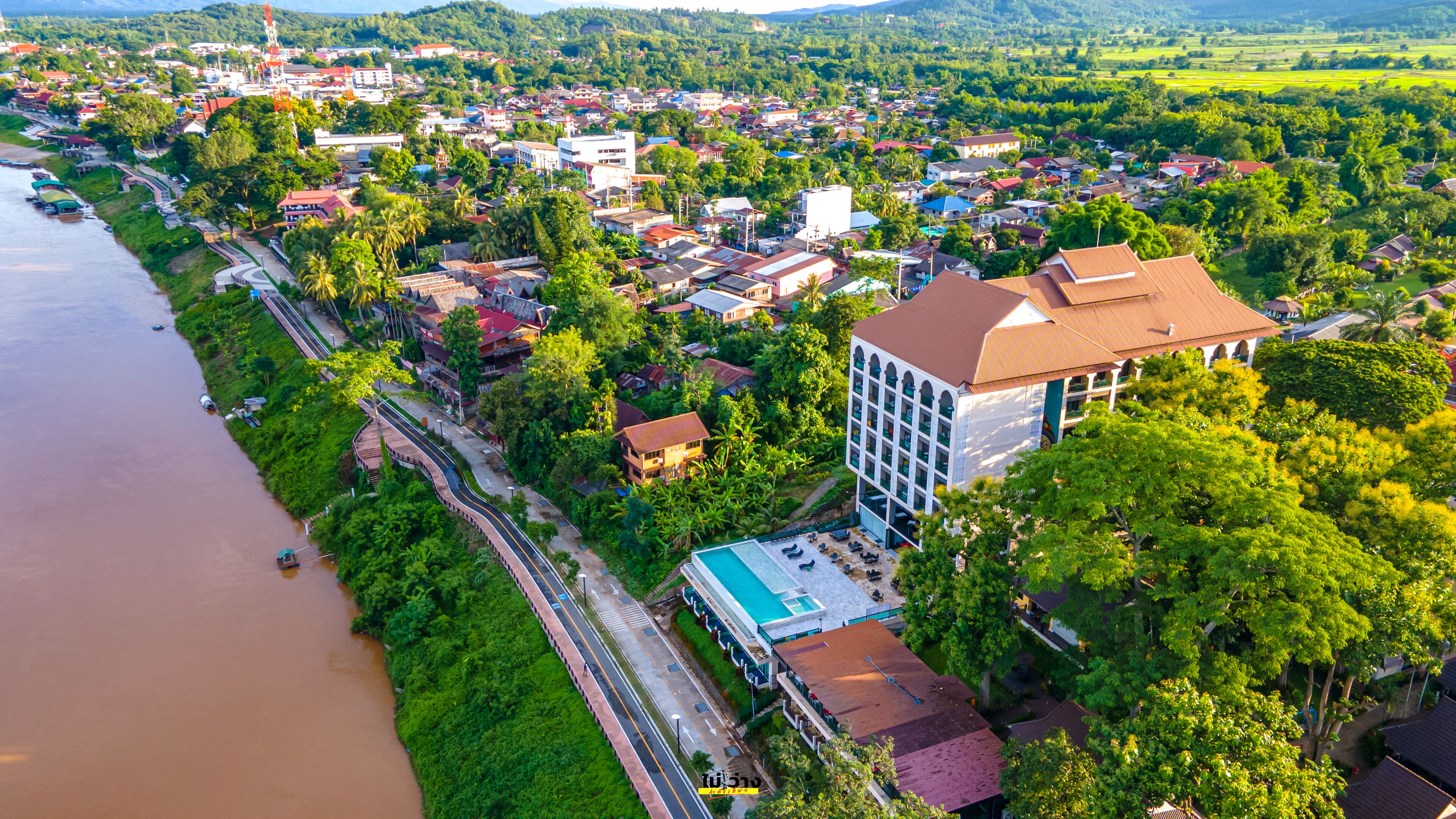 Une vue aérienne d’une ville côtière, avec un hôtel imposant doté d’une piscine, entouré d’une végétation luxuriante et bordé par une rivière.