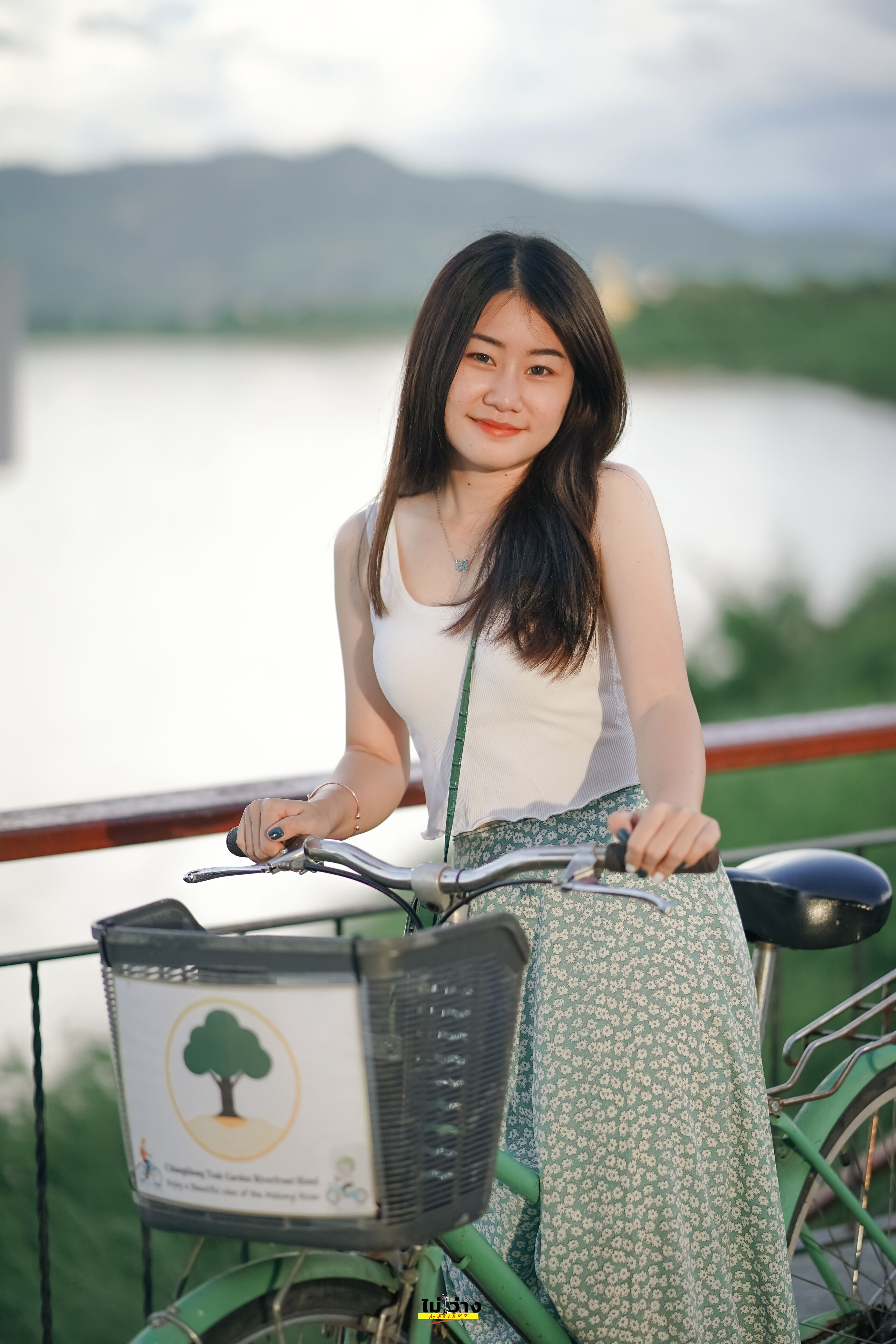A person sitting on a green bicycle with a basket, near a body of water and a railing.