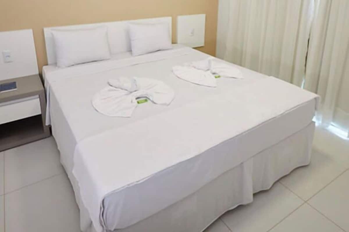 A hotel room with a large bed, white bedding, a nightstand, and a window with curtains.
