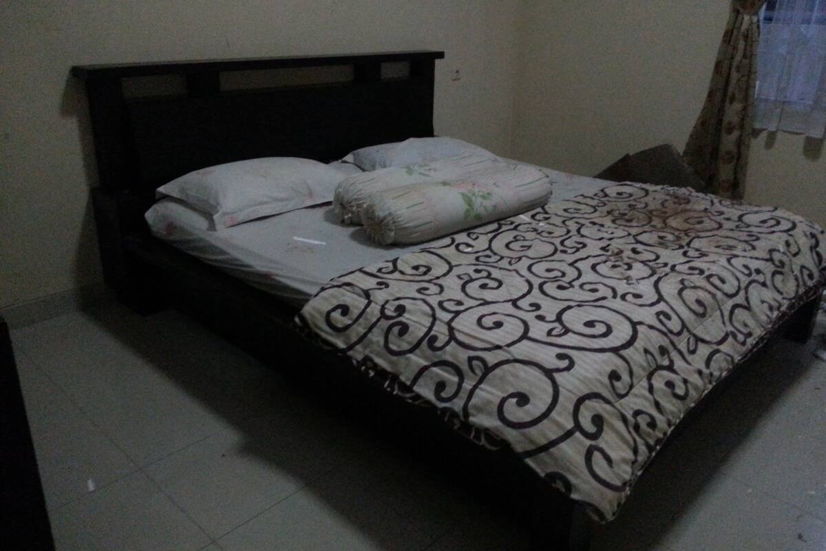 Una cama individual con una colcha estampada y almohadas, cabecera oscura y pared lisa con cortina.
