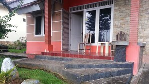 A house with a red and beige exterior, a stone staircase leading to a door, and a small garden area with rocks and plants. - Kota Bunga C (Cipanas)