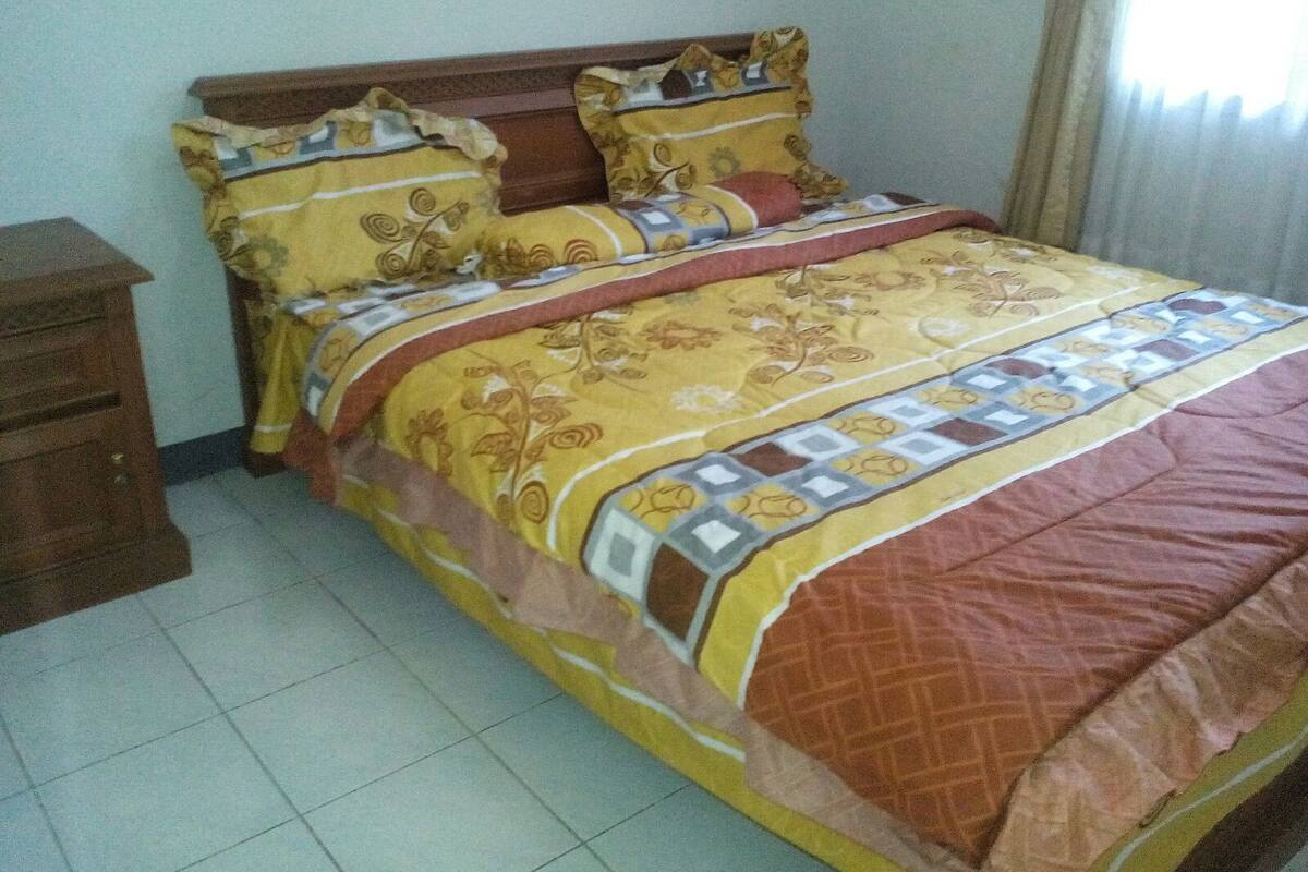 Una cama bien hecha con una colcha estampada y almohadas, una mesita de noche de madera a la izquierda, y una ventana con cortinas a la derecha.