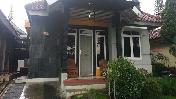 A two-story house with a sloped roof, a front porch with a bench, and a small garden area. - Kota Bunga C (Cipanas)