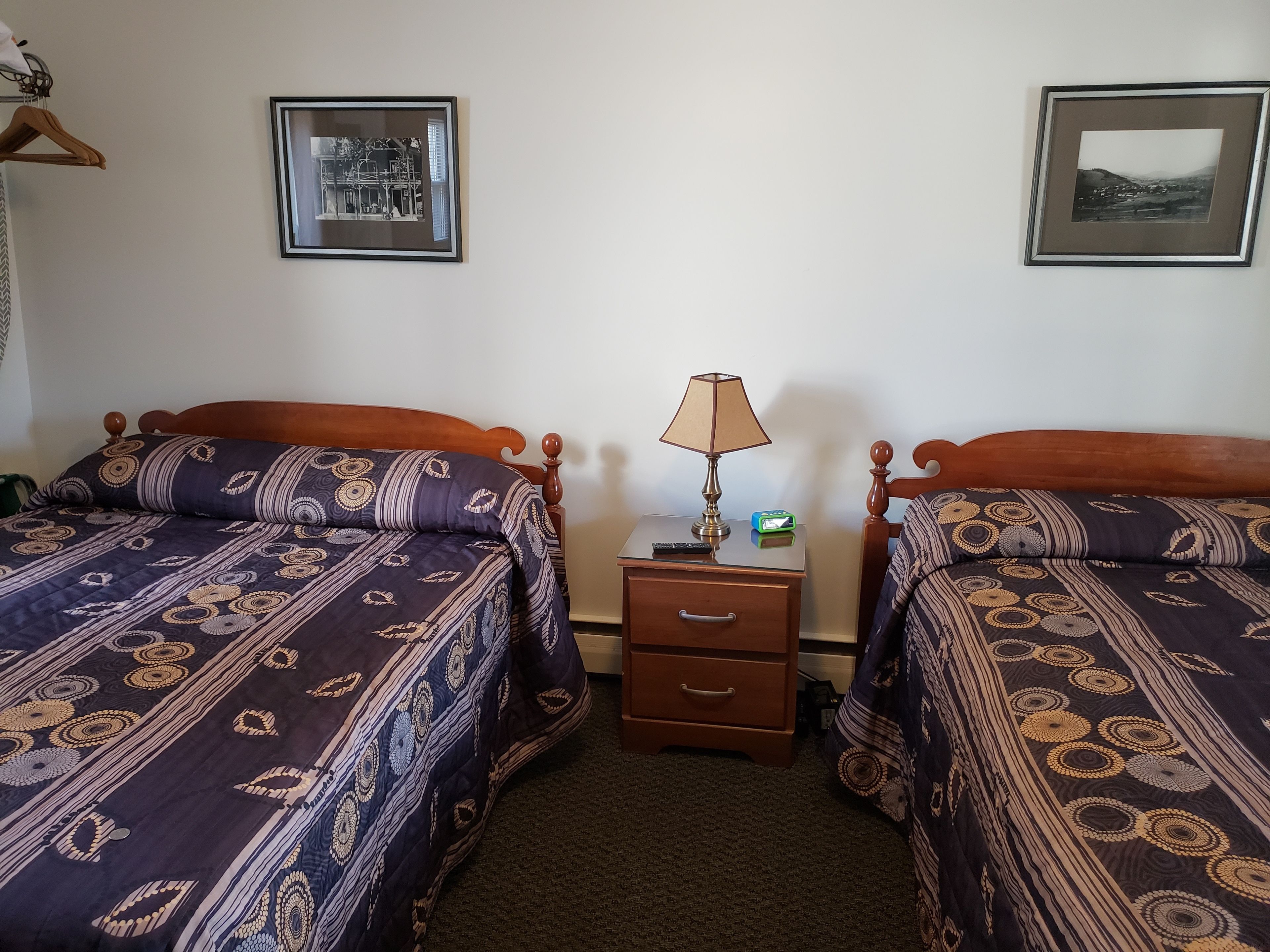 Habitación de hotel con dos camas, una mesita de noche con lámpara y reloj, y dos cuadros en la pared.