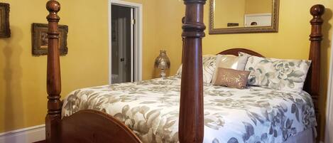 A four-poster bed with floral bedding, a mirror above it, and a door leading to another room.