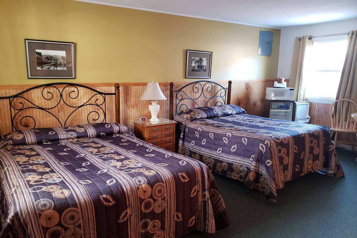 Une chambre d’hôtel avec deux lits, des têtes de lit en bois, une table de chevet avec une lampe, une commode et une fenêtre avec des rideaux.