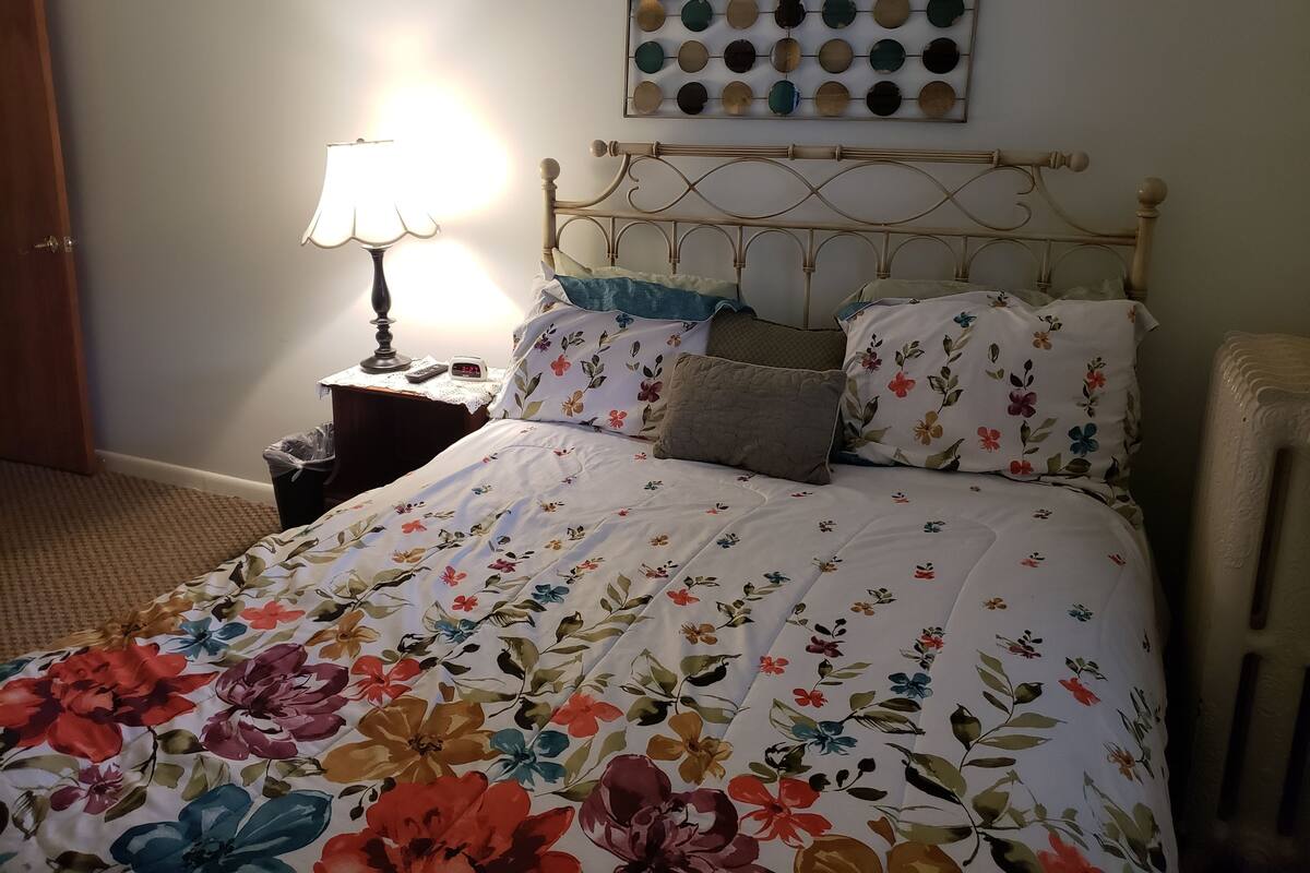 Un lit bien fait, avec une literie à motifs floraux, une lampe de chevet, une horloge et un tableau décoratif au mur.