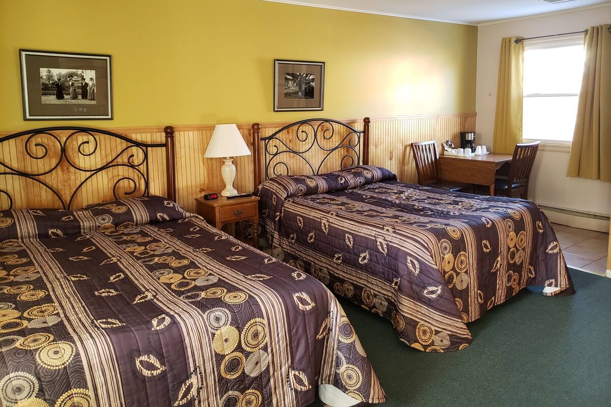 Une chambre d’hôtel avec deux lits, une table de chevet avec une lampe, un bureau avec une chaise et deux tableaux encadrés au mur.