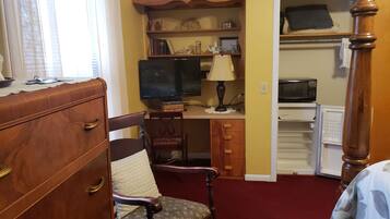 A room with a wooden dresser, a chair with a cushion, a desk with a computer and lamp, a television, and a closet with a microwave.
