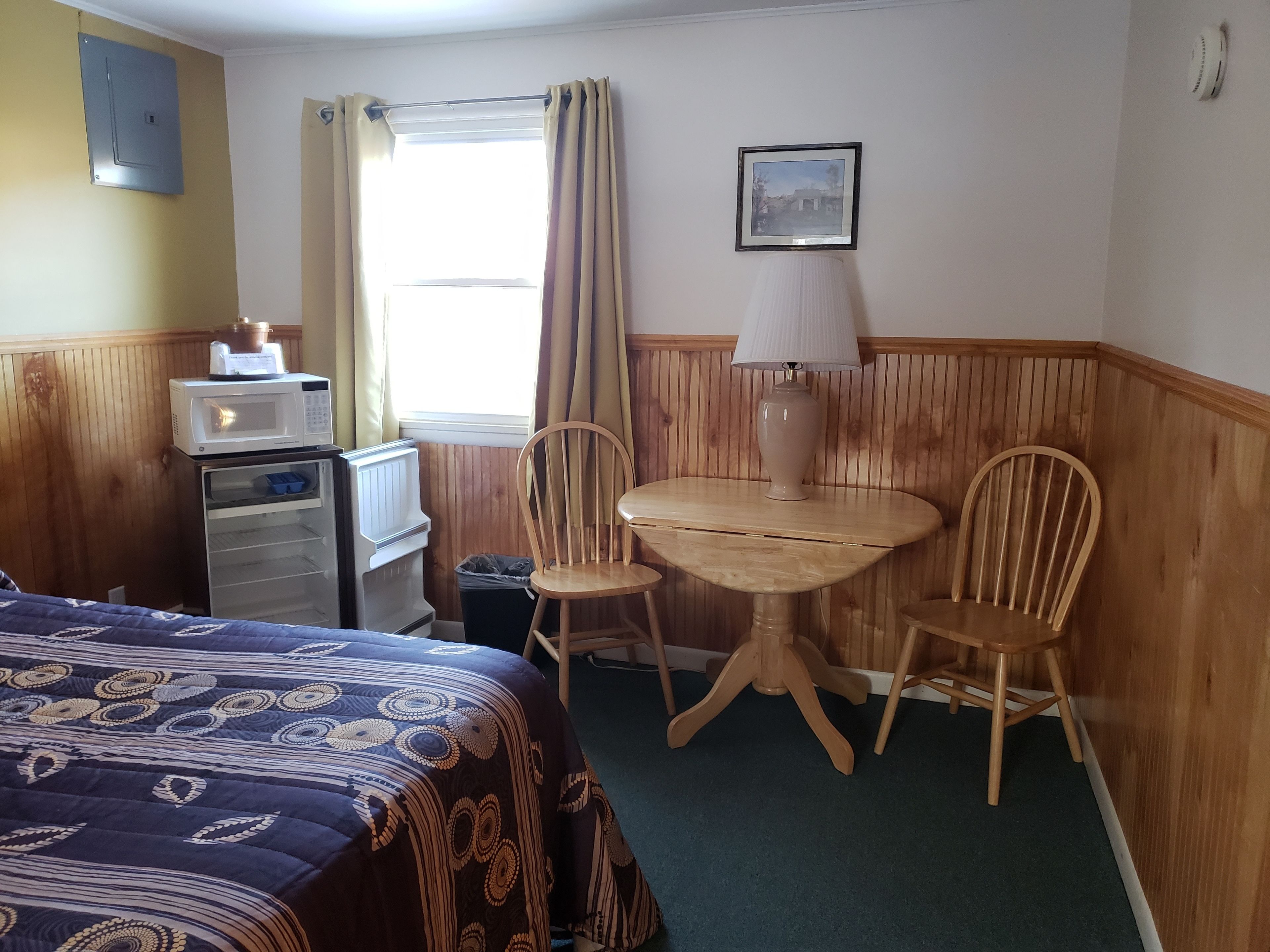 A hotel room with a bed, a small desk with a microwave, a chair, a table with a lamp, a picture on the wall, and a window with curtains.