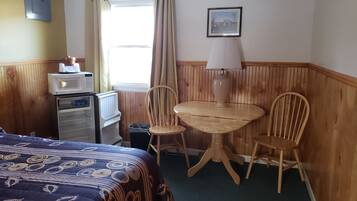 A hotel room with a bed, a small desk with a microwave, a chair, a table with a lamp, a picture on the wall, and a window with curtains.