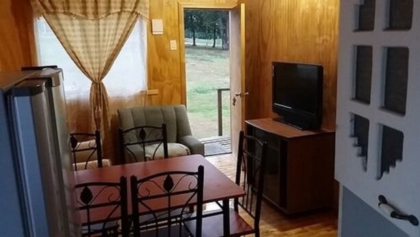 Una habitación pequeña con puerta de madera, un televisor sobre un mueble bajo, una mesa de comedor con sillas y vistas a un espacio exterior con césped.