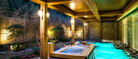 A heated outdoor pool area with wooden beams and stone walls, surrounded by a garden with various plants and a stone wall.