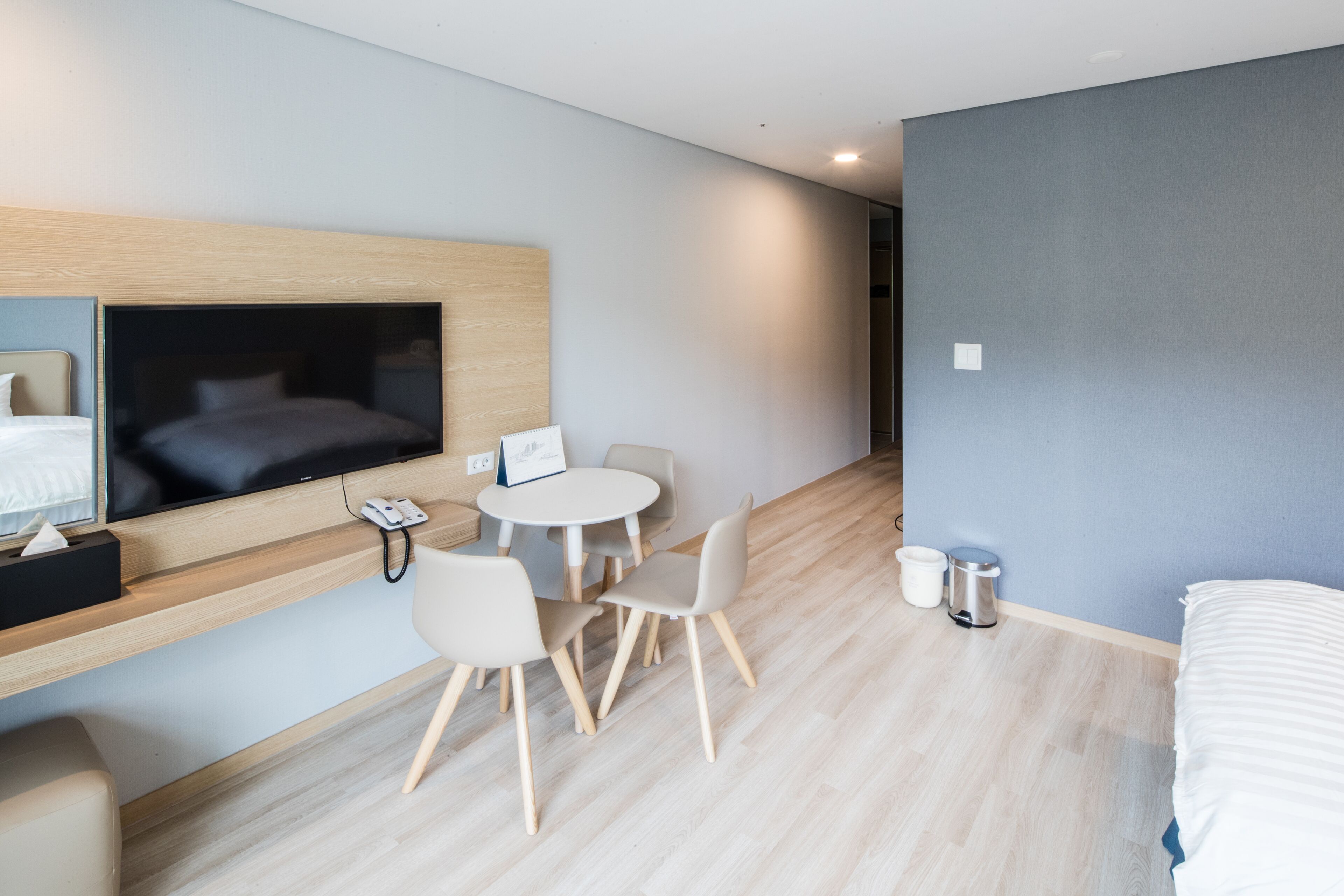 A modern hotel room with a bed, a TV mounted on the wall, a desk with a chair, a small table with chairs, and a trash bin.
