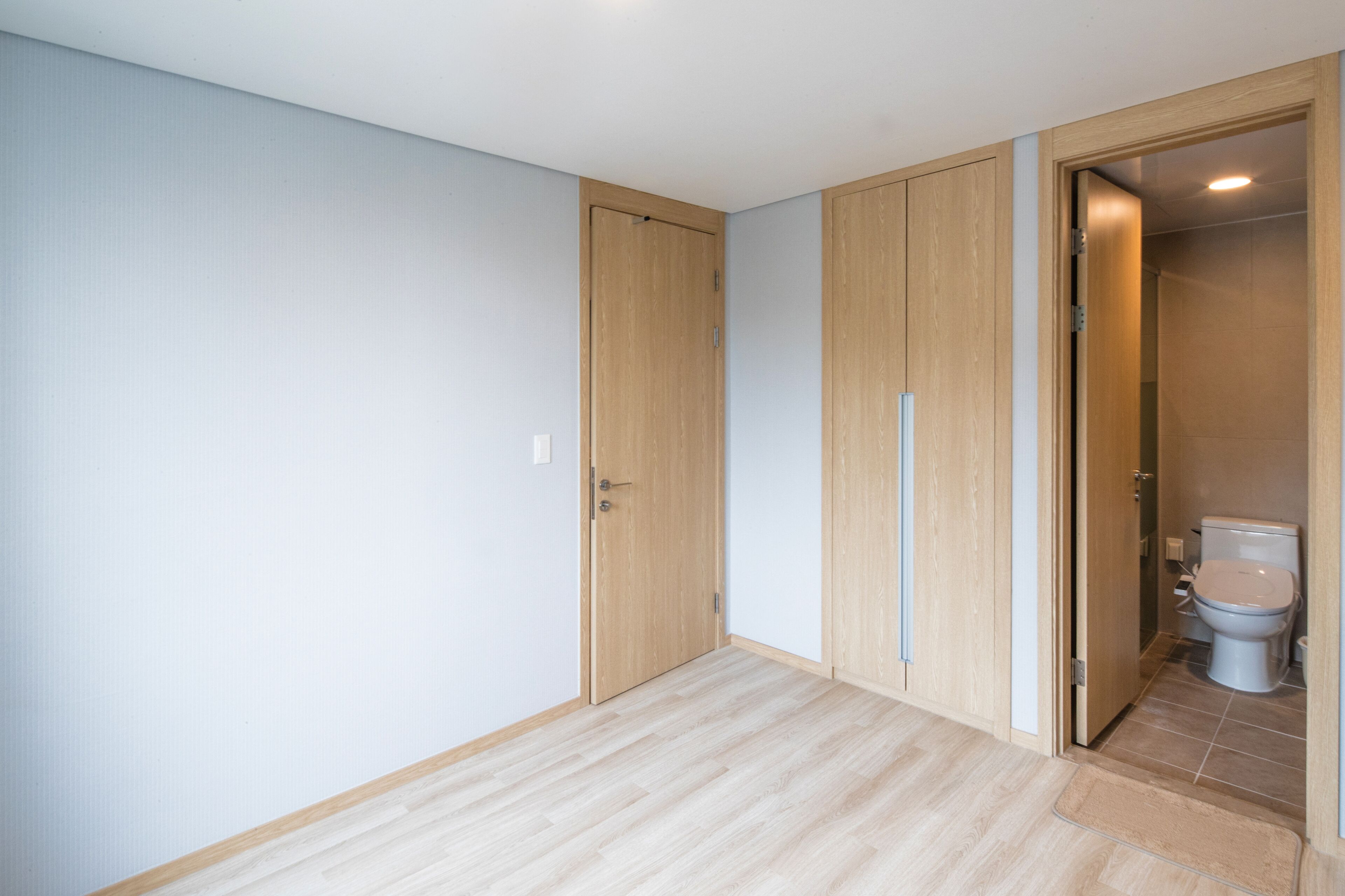 Una habitación con puerta de madera, piso de tonos claros y un baño contiguo donde se ve un inodoro.