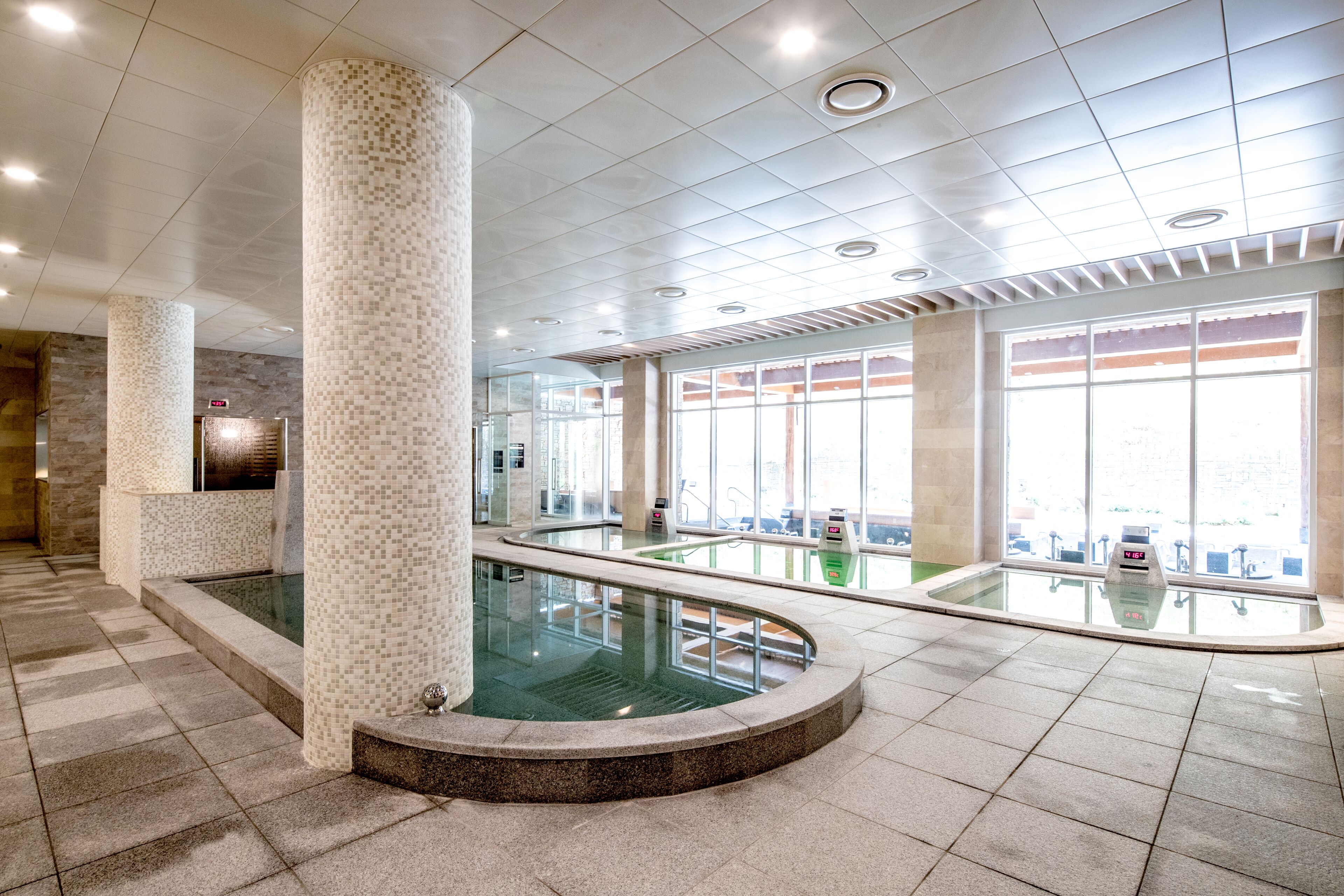 Uma área interna de piscina moderna com pisos azulejados, guarda-corpos de vidro e grandes janelas.